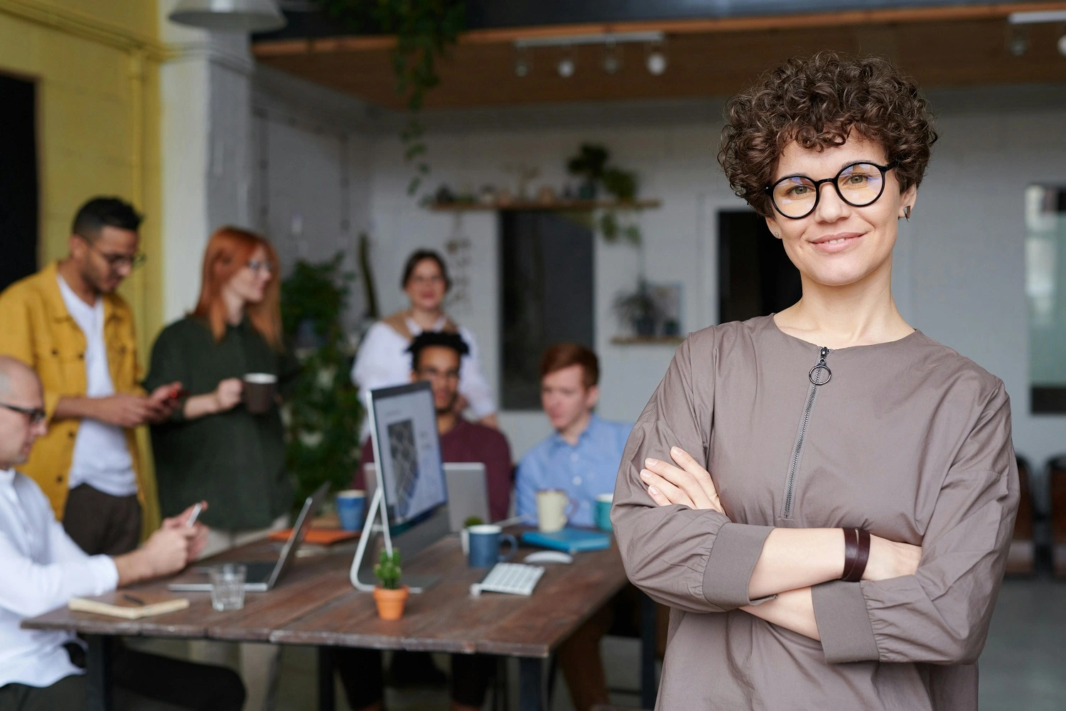 Uma mulher sorrindo de braços cruzados com um grupo de pessoas trabalhando ao fundo, ilustrando o conceito de ações de liderança.